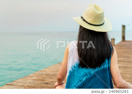 Rear view of a woman wearing a sun hat standing on a wooden pier overlooking calm sea. Travel, freedom, peaceful vacation, tropical destination, relaxation, wellness. Tropical beach destination. Rear view of a woman wearing a sun hat standing on a wooden pier overlooking calm sea. Travel, freedom, peaceful vacation, tropical destination, relaxation, wellness. Tropical beach destination. 140983261