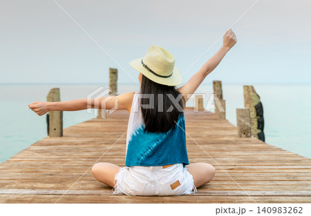 Rear view of a happy woman stretching her arms while sitting on a wooden pier overlooking calm sea. Freedom, travel, peaceful vacation, tropical lifestyle, wellness, relaxation. Summer holiday. Rear view of a happy woman stretching her arms while sitting on a wooden pier overlooking calm sea. Freedom, travel, peaceful vacation, tropical lifestyle, wellness, relaxation. Summer holiday. 140983262