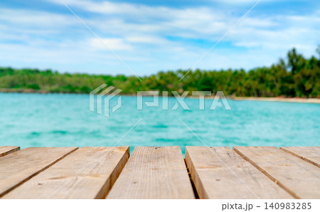 Empty wooden tabletop on a tropical beach pier overlooking turquoise ocean with soft blurred coastline and blue sky. Natural product display background for travel, summer, advertising, and branding. Empty wooden tabletop on a tropical beach pier overlooking turquoise ocean with soft blurred coastline and blue sky. Natural product display background for travel, summer, advertising, and branding. 140983285