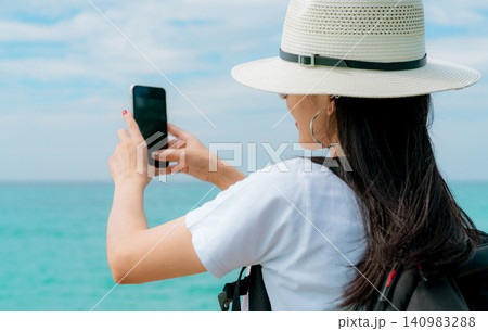 A tourist photographing tropical coastline with a mobile phone. Modern travel, digital storytelling, seaside relaxation, and unforgettable holiday experiences. Summer vacation lifestyle, tourism. A tourist photographing tropical coastline with a mobile phone. Modern travel, digital storytelling, seaside relaxation, and unforgettable holiday experiences. Summer vacation lifestyle, tourism. 140983288