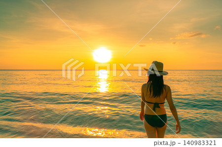 Back view of a woman in a bikini and sun hat standing at the shoreline while watching tropical sunset over calm ocean. Summer vacation, beach travel, luxury resort, relaxation. Summer holiday. Back view of a woman in a bikini and sun hat standing at the shoreline while watching tropical sunset over calm ocean. Summer vacation, beach travel, luxury resort, relaxation. Summer holiday. 140983321