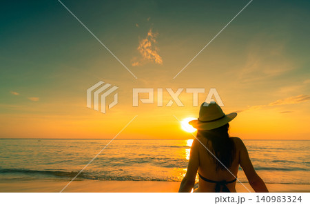 Woman wearing sun hat enjoying tranquil tropical sunset while sitting on beach facing ocean. Luxury vacation, island tourism, golden hour, seaside serenity, relaxation, and inspiring travel lifestyle. Woman wearing sun hat enjoying tranquil tropical sunset while sitting on beach facing ocean. Luxury vacation, island tourism, golden hour, seaside serenity, relaxation, and inspiring travel lifestyle. 140983324