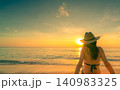 Woman traveler relaxing on sandy shore during a breathtaking golden sunset beside tropical sea. Peaceful ocean reflections, summer holiday, beach destination, luxury resort, and mindful escape concept 140983325