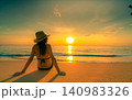 Back view of a woman in a bikini and sun hat sitting on a tropical beach while watching sunset over the calm ocean. Summer vacation, seaside relaxation, luxury travel, wellness, and coastal lifestyle. 140983326