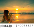 Woman relaxing on a tropical sandy beach while watching sunset over calm ocean. Luxury summer vacation. Island getaway, luxury resort, oceanfront relaxation, wellness, and coastal travel experience. 140983327