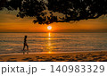 Silhouette of woman walking along a tropical beach at sunset. Golden hour travel, summer vacation, coastal wellness. Peaceful holiday, wellness travel, leisure lifestyle, and summer destination. 140983329