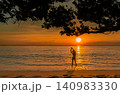 Silhouette of woman stretching in gentle ocean waves beneath a tropical tree at golden sunset. Peaceful beach wellness, summer travel, relaxation, coastal lifestyle, and serene vacation destination. 140983330