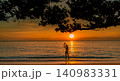 Silhouette of woman stretching in gentle ocean waves beneath a tropical tree at golden sunset. Peaceful beach wellness, summer travel, relaxation, coastal lifestyle, and serene vacation destination. 140983331