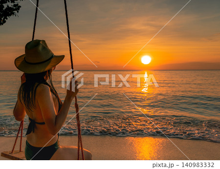 Golden sunset reflecting across the tropical ocean as a woman enjoys beach swing beside gentle waves. Luxury vacation, serene coastal destination, summer travel, relaxation, mindfulness, and wellness. 140983332