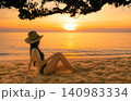 Golden sunset over a tranquil tropical shoreline with woman relaxing on sand under seaside tree. Beach vacation, luxury travel, wellness retreat, summer lifestyle, ocean horizon, and coastal paradise. 140983334