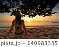 Golden sunset over tropical shoreline with woman relaxing on the sand under a seaside tree. Beach vacation, luxury travel, wellness retreat, summer lifestyle, ocean horizon, and coastal paradise. 140983335