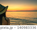 Close up woman in sun hat watching a glowing tropical ocean sunset. Peaceful beach relaxation, summer vacation, wellness lifestyle, golden hour travel, tranquil seaside escape, and coastal serenity. 140983336