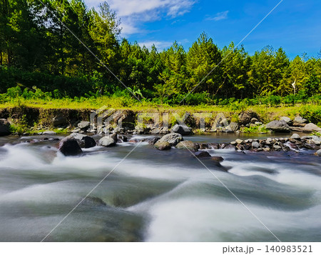 Scenic long exposure landscape photograph of a rocky river with silky smooth water flowing through a lush green valley in Songgon, Banyuwangi, Indonesia. Densely packed pine trees and tropical foliage Scenic long exposure landscape photograph of a rocky river with silky smooth water flowing through a lush green valley in Songgon, Banyuwangi, Indonesia. Densely packed pine trees and tropical foliage 140983521