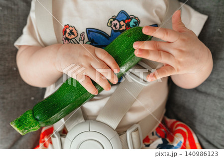 Child holding zucchini, in indoor possibly home, with zucchini, car seat, with plastic, fabric texture, from a close-up top-down perspective perspective, with green, white accents. Child holding zucchini, in indoor possibly home, with zucchini, car seat, with plastic, fabric texture, from a close-up top-down perspective perspective, with green, white accents. 140986123