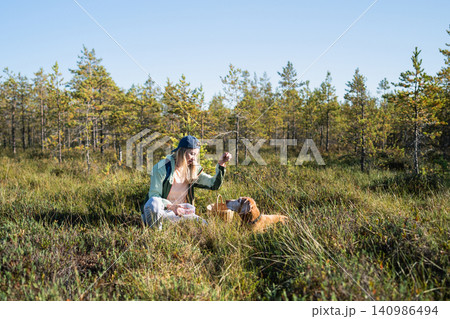 Happy woman hiker collects fresh edible berries sitting at marsh and playing with beloved Vizsla dog Happy woman hiker collects fresh edible berries sitting at marsh and playing with beloved Vizsla dog 140986494