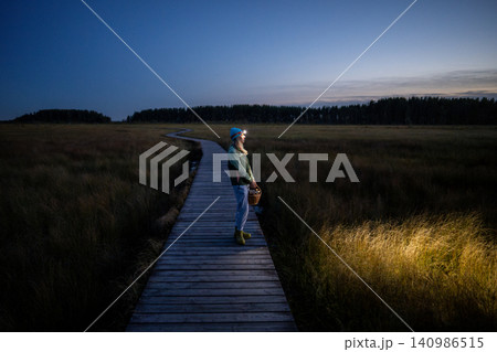Woman nature lover enjoys autumn sunset while standing on wooden path with basket full of mushrooms. Woman nature lover enjoys autumn sunset while standing on wooden path with basket full of mushrooms. 140986515