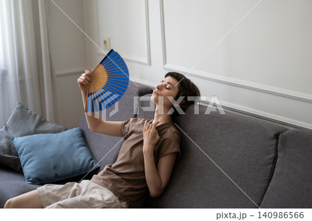 Exhausted sweaty woman fanning herself sitting on sofa suffering from summer heat without AC Exhausted sweaty woman fanning herself sitting on sofa suffering from summer heat without AC 140986566