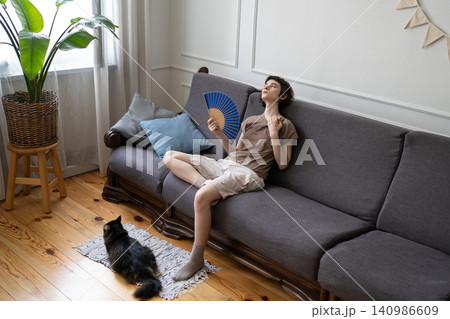 Sweaty woman using fan for relief, cat in heat exhaustion nearby. Home climate struggle, no AC Sweaty woman using fan for relief, cat in heat exhaustion nearby. Home climate struggle, no AC 140986609