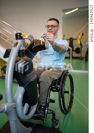 Serious disabled man in wheelchair using hand cycling machine in gym for recovery after injury. 140987025