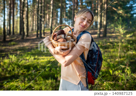 Pleased relaxed woman forager on mushroom picking in forest standing hugging basket full of porcini Pleased relaxed woman forager on mushroom picking in forest standing hugging basket full of porcini 140987125