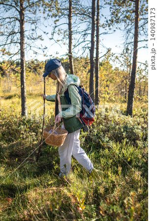 Focused relaxed woman forager with basket making way through wetlands helping herself with stick Focused relaxed woman forager with basket making way through wetlands helping herself with stick 140987173
