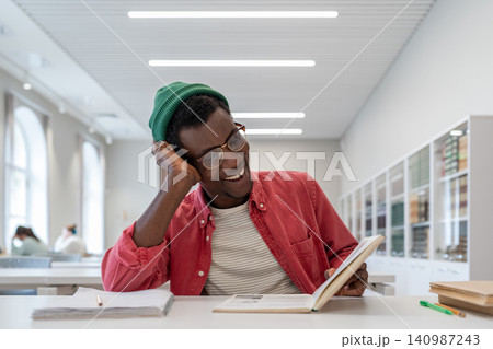 Laughing cheerful ethnic African American man reading book sits at table during break in college Laughing cheerful ethnic African American man reading book sits at table during break in college 140987243