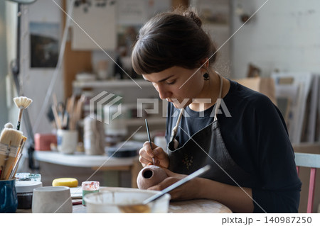 Lady master paints clay jug with serious and concentrated expression sitting at table in workshop Lady master paints clay jug with serious and concentrated expression sitting at table in workshop 140987250