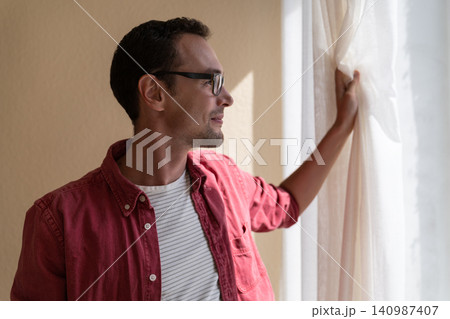 Smiling German man holding on to curtain and looking out window in summer weather admiring cityscape 140987407