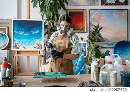 Woman in respirator mixing varnish and epoxy in cup at workshop table, working process in gallery. Woman in respirator mixing varnish and epoxy in cup at workshop table, working process in gallery. 140987456