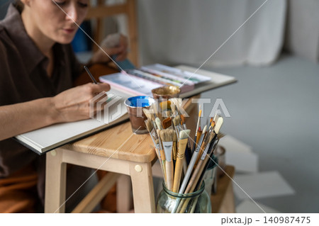 Serious focused woman painter drawing pictures at home on chair using watercolors and sketchbook. Serious focused woman painter drawing pictures at home on chair using watercolors and sketchbook. 140987475