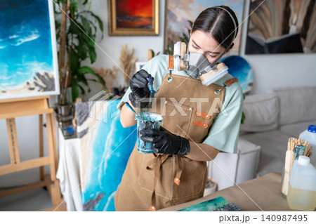 Talented woman artist creating liquid art, holding cup and dripping colored pigment into varnish. Talented woman artist creating liquid art, holding cup and dripping colored pigment into varnish. 140987495