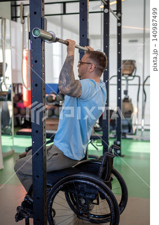 Man in wheelchair doing vertical pull ups workout using adaptive fitness equipment in sports center. 140987499