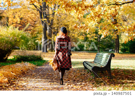 redhead woman in plaid coat and beret walks through the park at sunny day, back view. fall season 140987501
