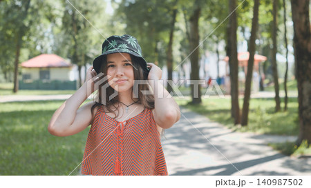 Young woman wearing trendy bucket hat, walking through sunlit park with lush greenery, basking in warm summer afternoon Young woman wearing trendy bucket hat, walking through sunlit park with lush greenery, basking in warm summer afternoon 140987502
