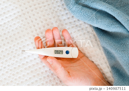 Closeup of man hand holding digital thermometer, lying on bed. High body temperature, flu Closeup of man hand holding digital thermometer, lying on bed. High body temperature, flu 140987511