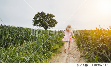 Young girl barefoot running across verdant meadow, reaching solitary tree during golden hour sunset lighting 140987512