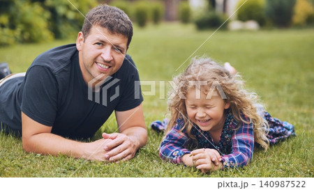 Smiling father bonding with young daughter, resting together on lush green lawn during relaxed family afternoon 140987522