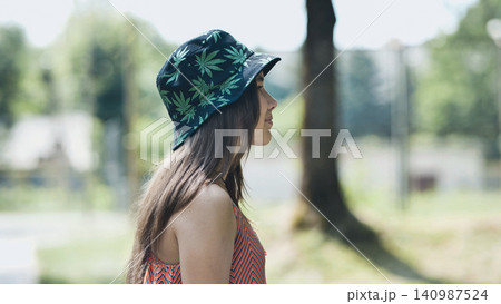 Young female relaxing in green park, wearing trendy bucket hat with marijuana leaf pattern, enjoying sunny afternoon Young female relaxing in green park, wearing trendy bucket hat with marijuana leaf pattern, enjoying sunny afternoon 140987524