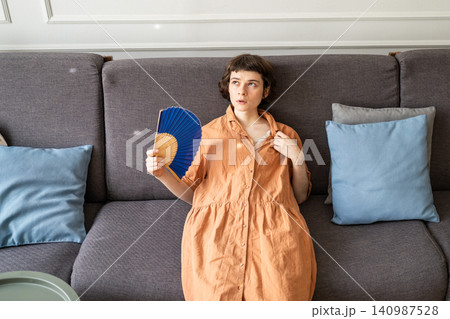 Woman on couch with sick expression waving hand fan overwhelmed by summer heat in apartment. 140987528