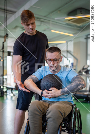Personal coach supporting disabled man in wheelchair during power workout with medicine ball in gym. 140987590