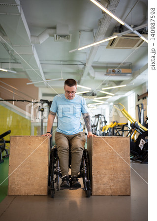 Confident man athlete in wheelchair doing push-ups strengthening exercise on wooden cubes in gym. Confident man athlete in wheelchair doing push-ups strengthening exercise on wooden cubes in gym. 140987598