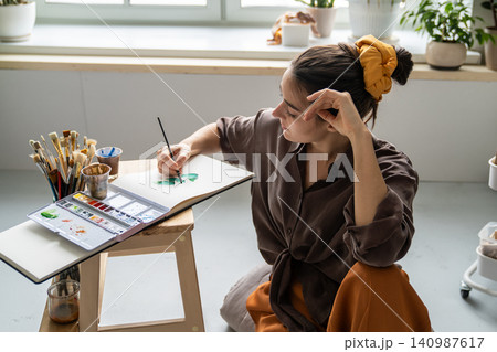 Talented woman spending free time with art hobby, painting pictures in sketchbook with aquarelle. Talented woman spending free time with art hobby, painting pictures in sketchbook with aquarelle. 140987617