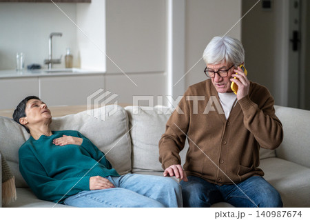 Senior woman lying on couch in room with pain in chest while husband talking to doctor on phone. Senior woman lying on couch in room with pain in chest while husband talking to doctor on phone. 140987674