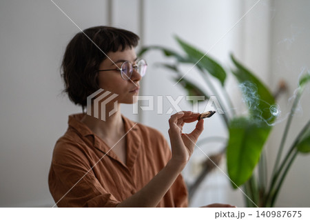 Relaxed woman practicing self care and mental health rituals, lightens palo santo stick in apartment Relaxed woman practicing self care and mental health rituals, lightens palo santo stick in apartment 140987675