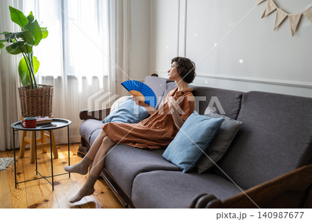 Tired woman suffers from overheating and stuffiness at home, sitting on sofa waving hand paper fan. Tired woman suffers from overheating and stuffiness at home, sitting on sofa waving hand paper fan. 140987677