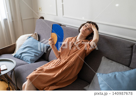 Woman lying on sofa in room fanning with paper fan suffering from head pain and summer heatstroke. 140987678