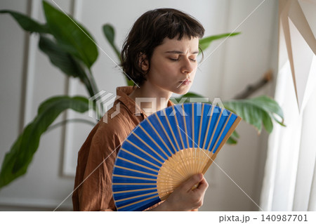 Woman at home annoyed by heatwave and heavy headache holding waving paper fan to cool down. Woman at home annoyed by heatwave and heavy headache holding waving paper fan to cool down. 140987701