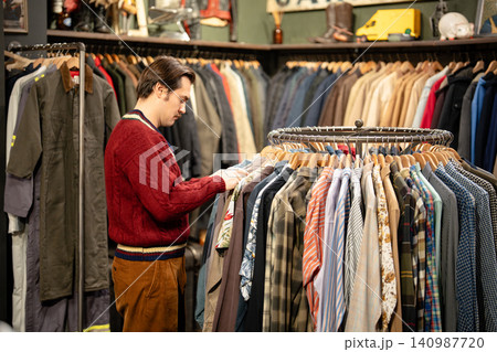 Handsome stylish man mindfully inspecting different shirts displayed on hanger in vintage shop. Handsome stylish man mindfully inspecting different shirts displayed on hanger in vintage shop. 140987720