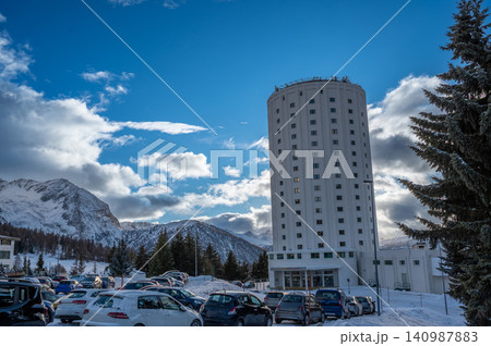 Sestriere, piedmont, italy. December 8, 2024. Sestriere, italy, presenting its iconic tower hotel, a bustling parking lot, and snow covered alps under a bright sky Sestriere, piedmont, italy. December 8, 2024. Sestriere, italy, presenting its iconic tower hotel, a bustling parking lot, and snow covered alps under a bright sky 140987883