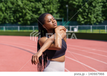 Black young woman with long braids stretches hands and shoulders standing on red track on stadium Black young woman with long braids stretches hands and shoulders standing on red track on stadium 140987966